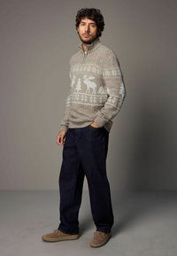 Beige gestrickter Pullover mit hohem Kragen, verziert mit blauen Mustern von Elchen und Bäumen; kombiniert mit dunkelblauen Hosen und hellbraunen Schuhen.