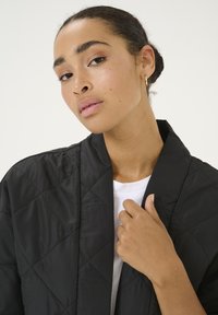 Sort quiltet jakke med diamantmønster, der har sjalskrave. Bæres med en hvid T-shirt underneath, uden mærkbare metaldele eller accenter.