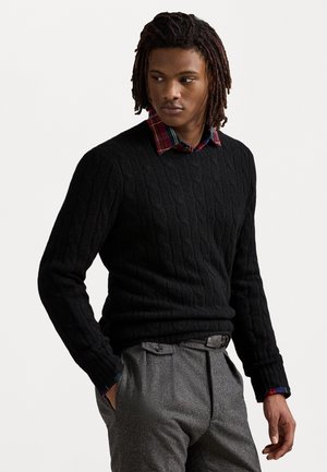 Maglione nero a trecce con collo rotondo, indossato sopra una camicia a quadri. Pantaloni grigi texture, mani nelle tasche, che mostrano un'accurata lavorazione.