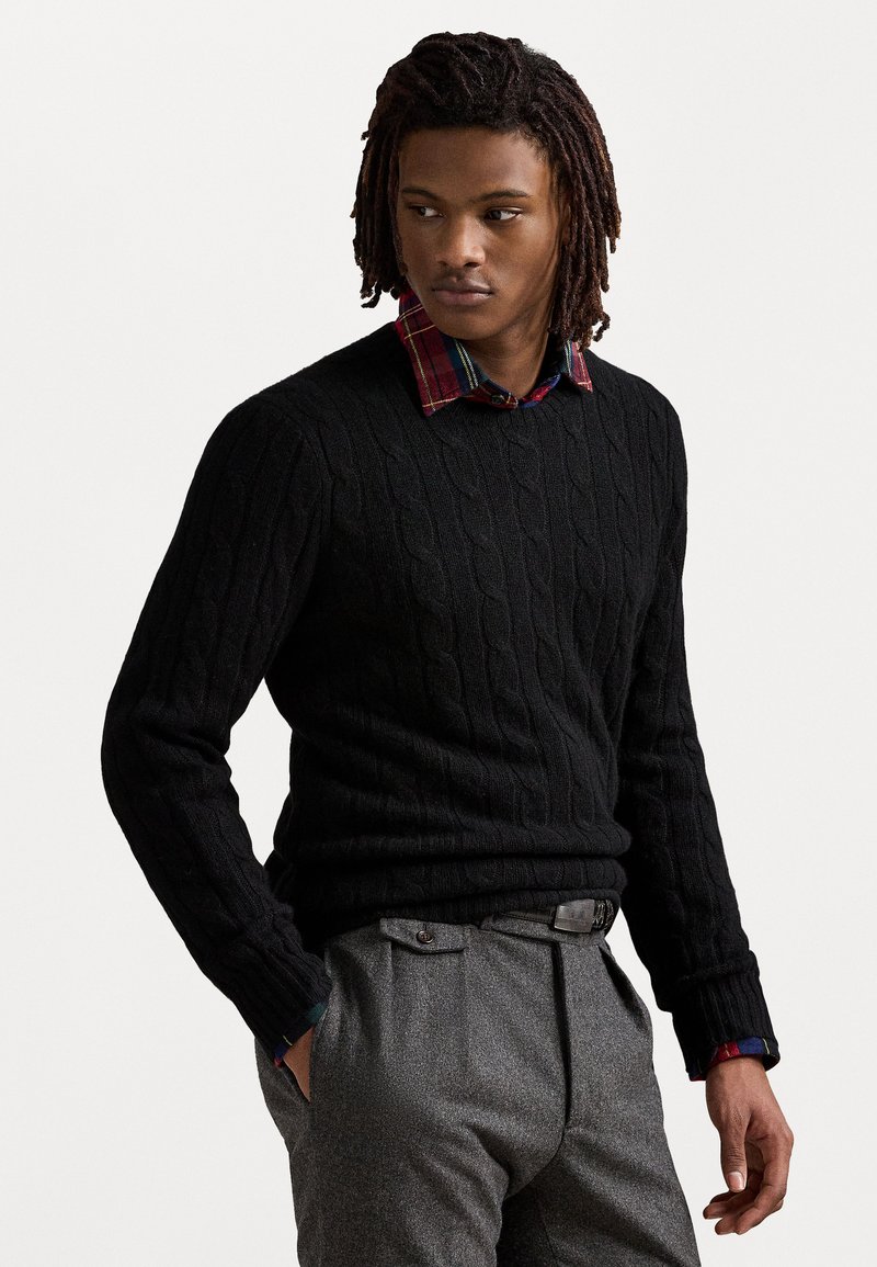 Μαύρος πλεκτός σκούφος με στρογγυλή λαιμόκοψη, φορεμένος πάνω από ένα καρό πουκάμισο. Γκρίζες υφασμένες παντελόνες, χέρια στις τσέπες, που δείχνουν λεπτομερή τεχνική κατασκευής.