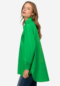 Camicia verde oversize con maniche lunghe, tasca frontale e design ricamato sul retro. Realizzata in tessuto morbido e leggero.