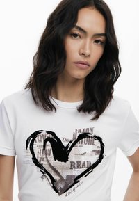 Weißes Baumwoll-T-Shirt mit einem großen schwarzen Herzmotiv, das Text und Zeitungsmotive zeigt. Weicher Stoff, runder Halsausschnitt, kurze Ärmel.