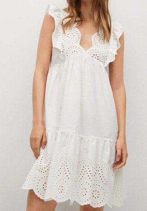 Nainen pukeutunut lyhyeen valkoiseen hihattomaan mekkoon, jossa on silmäreikäkirjailua, röyhelöiset olkapäät ja aaltoileva helmaversio, seisoo yksiväristä taustaa vasten.