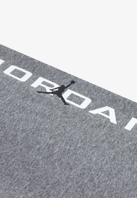 Un tissu gris avec une texture subtile présente un logo blanc "JORDAN" et une silhouette noire d'une figure en train de sauter, indiquant un design de vêtements de sport.