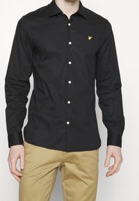 Uomo che indossa una camicia nera aderente con un logo giallo ricamato sul petto e pantaloni beige, in piedi contro uno sfondo neutro.