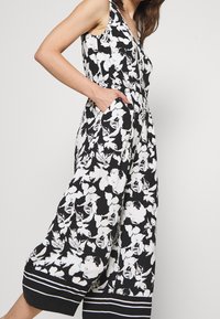 Jumpsuit floral en blanco y negro con escote en V, diseño sin mangas, bolsillos y dobladillo a rayas. Tejido ligero con una textura suave.