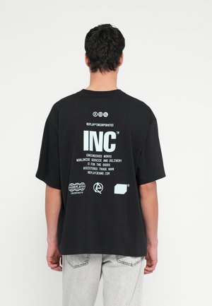 Mies selinpäin, musta oversize-t-paita, jossa valkoista painettua tekstiä ja logoja, yhdistetty vaaleanharmaisiin farkkuihin, yksinkertaisen taustan edessä.