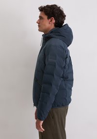 Marineblauwe donsjas met capuchon, voorzien van een ritssluiting aan de voorkant en een gestructureerde afwerking. Heeft een patch op de mouw en elastische manchetten.