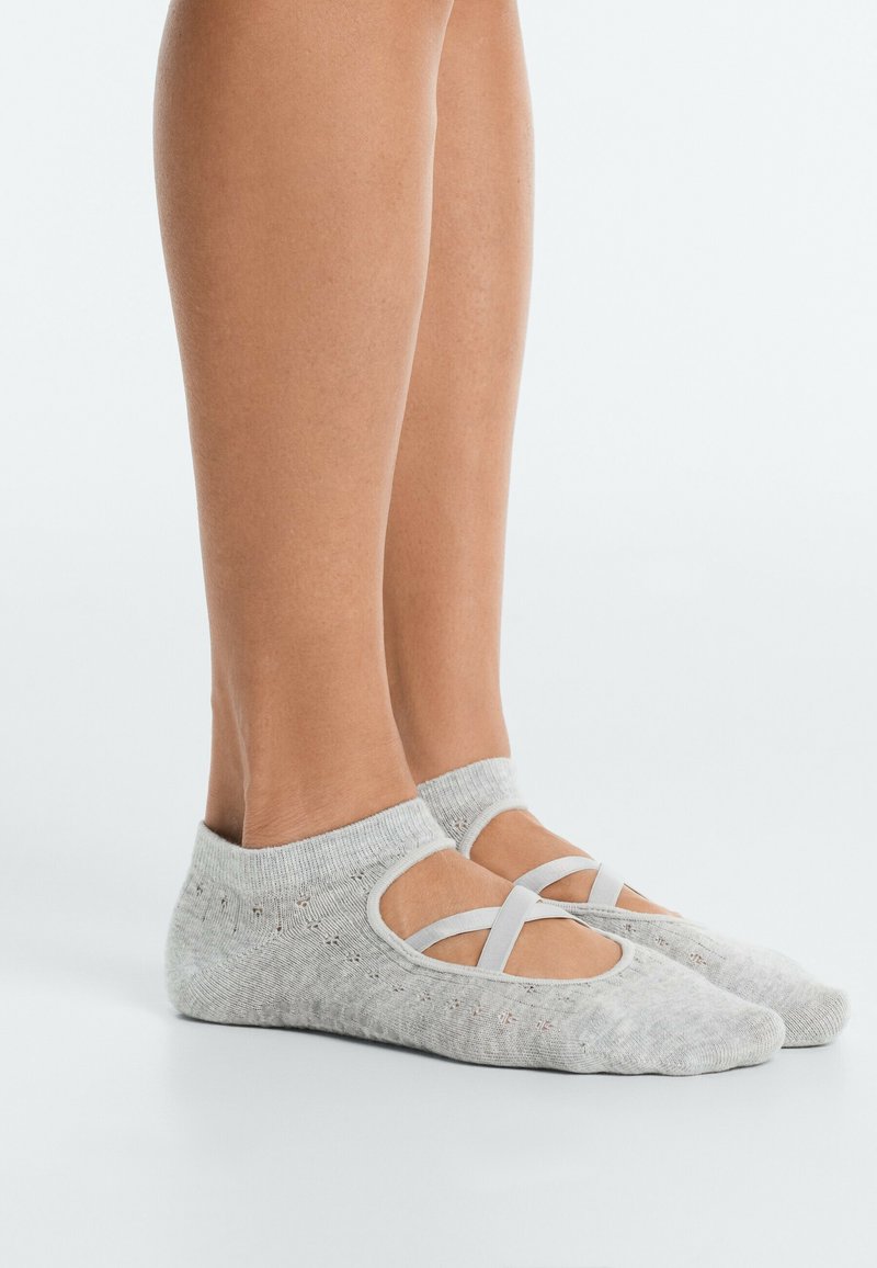 Jambes portant des chaussettes grises basses avec des brides croisées sur le dessus, debout sur une surface blanche avec un fond uni.