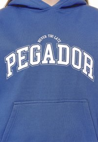 Modrá mikina s kapucí a bílým textem "PEGADOR" velkými písmeny, s menším textem "NEVER TOO LATE" nad hlavním textem, viditelná přední kapsa ve stylu klokan.