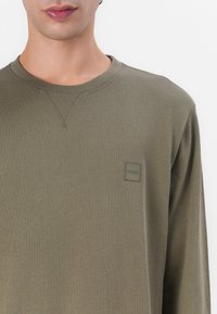 Olivengrønn sweatshirt har rund hals og lange ermer, med et lite rektangulært logo-merke i en matchende tone på brystet.