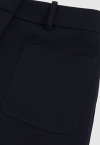 Gros plan d’un pantalon noir montrant une poche arrière et un passant de ceinture avec des coutures détaillées sur le tissu.