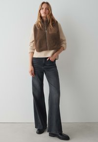 Gilet en fausse fourrure brune avec une patch sur la poitrine, pull en tricot beige, jean noir évasé, et chaussures noires sur fond clair.