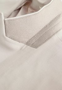 Helles beiges Gewebe mit einem strukturierten Kragen, das ein mehrschichtiges Design aufweist und an den Kanten sichtbare Nahtdetails besitzt.