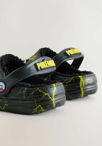 Zwarte Pokémon-slippers met een gele bliksempatroon, zachte binnenvoering, verstelbare band en groot geel logo aan de voorkant.