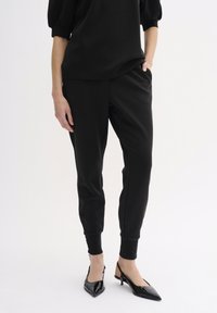Schwarze Trainingshose mit lockerer Passform, elastischen Bündchen, seitlichen Taschen, kombiniert mit spitzen schwarzen Slingback-Highheels. Weicher Stoff.