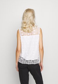 Vit spetsärmlös topp med intrikata mönster och en scalloped fåll, parat med svarta byxor. Bakifrån visas den detaljerade spetsdesignen.