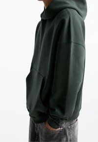 Felpa verde scuro con una tasca frontale ampia, polsini a coste e cappuccio con coulisse, realizzata in morbido materiale di peso medio.