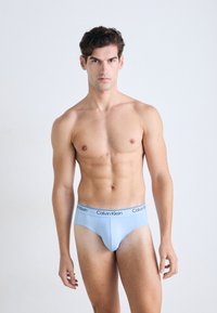 Junger Mann mit athletischem Körperbau, der vor einem schlichten Hintergrund steht, leichte blaue Calvin Klein Unterwäsche trägt und mit neutralem Ausdruck geradeaus schaut.
