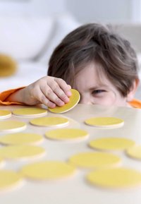 Holzspielsteine mit einer glatten, matten gelben Oberfläche, kreisförmiger Form, verstreut auf einem hellen Holztisch, während eine Kinderhand einen aufnimmt.