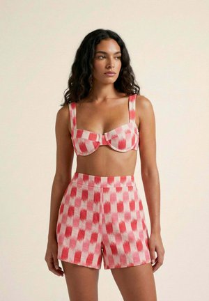 Mujer con cabello largo y rizado vistiendo un top corto de cuadros rojos y rosas con shorts a juego de talle alto, posando frente a un fondo liso.