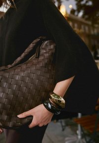 Brown woven leather bag with a slouchy shape and zipper closure, paired with stacked bangles in gold, black, and brown tones.