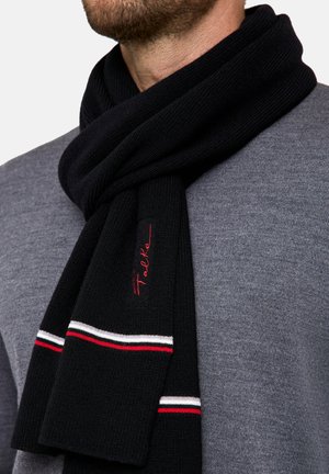 Écharpe noire en tricot avec des rayures rouge, blanche et rouge. Un petit logo est brodé en rouge. Portée par-dessus un pull gris.