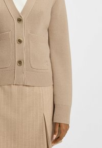 Beżowy sweter kardigan z dekoltem w kształcie litery V, czterema guzikami i dwiema przednimi kieszeniami. Żebrowane mankiety i dół. Teksturowana tkanina, subtelna marka na kieszeni.