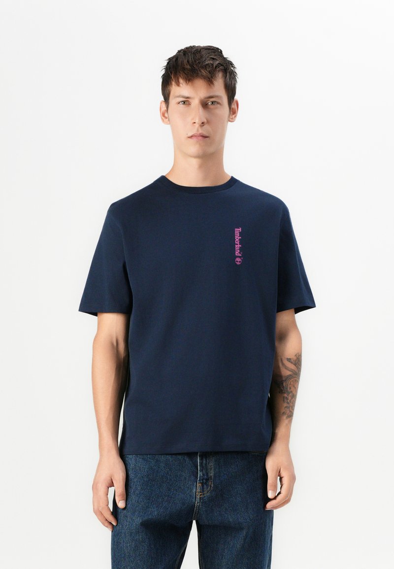 Młody mężczyzna w granatowej koszulce Timberland i niebieskich dżinsach, stojący na tle białej ściany, prezentujący tatuaż na lewej przedramieniu.