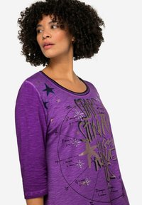 Lila Langarmshirt mit Rundhalsausschnitt, das mit metallischem Text und Sternmustern versehen ist. Leichter Stoff mit einer strukturierten Optik.