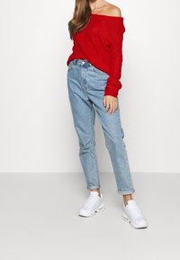 Roter Strickpullover mit schulterfreier Form, hellblaue High-Waist-Jeans und weiße Sneakers. Lässiger Schnitt mit geripptem Muster und hochgerollten Säumen.