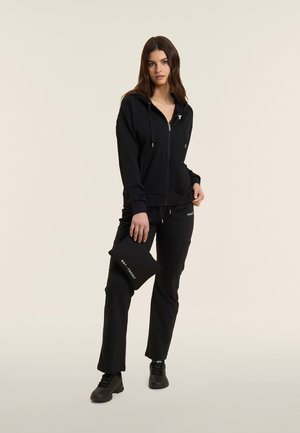 Donna in piedi con felpa nera con zip e pantaloni, che tiene una pochette nera con testo bianco, su uno sfondo chiaro uniforme.