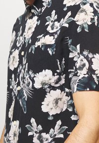 Zbliżenie osoby w czarnej koszulce z krótkim rękawem z białym kwiatowym wzorem i jasnymi żółtymi guzikami.