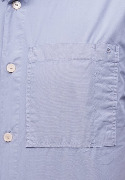 Φωτεινό γαλάζιο πουκάμισο με μεγάλη τετράγωνη τσέπη στο στήθος με ζιγκ-ζαγκ ραφές και κρεμ κουμπιά στο μπροστινό μέρος.