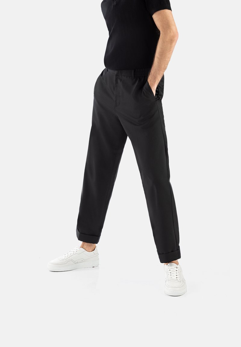 Pantalons noirs tapered avec une taille élastique, poches latérales et ourlets roulés, assortis à des baskets blanches sur un fond neutre.