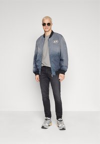 Veste bomber bleue, T-shirt gris, jeans noirs et baskets grises avec des accents orange. La veste présente un logo rond sur la poitrine gauche.