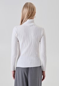 Donna con lunghi capelli biondi che indossa un maglione bianco a collo alto testurizzato e pantaloni grigi, vista di spalle su uno sfondo neutro.