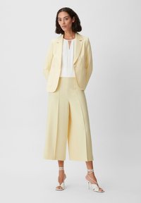 Frau, die in einem cremfarbenen Blazer und weit geschnittenen, cropped Hosen mit einer weißen Bluse und weißen High Heels vor einem schlichten Hintergrund steht.