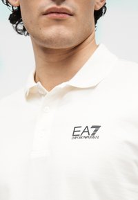 Polo branca feita de algodão, com gola clássica e pala com dois botões. Apresenta o logótipo EA7 Emporio Armani no peito.