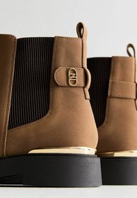 Botines de ante marrón con paneles de pana negra, detalles en dorado y suela de goma negra. Presenta una hebilla decorativa y un tirador.