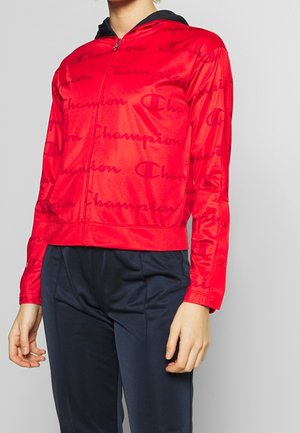 Personne portant un sweat à capuche zippé rouge avec un motif logo "Champion" et un pantalon bleu marine, cadrée des épaules jusqu'aux hanches.