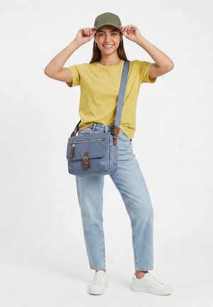 Smilende kvinde, der justerer en grøn kasket, iført en gul t-shirt, lyseblå jeans, hvide sneakers og bærer en blå crossbody-taske.
