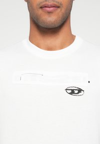 Λευκό βαμβακερό t-shirt με μαύρο γραφικό μάτι και μερικώς καλυμμένο κείμενο μπροστά. Κλασικός στρογγυλός λαιμός και κοντά μανίκια.