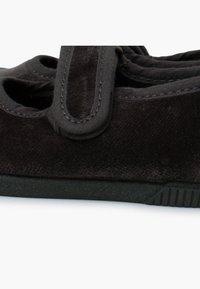 Zapato de ante negro con forma redondeada, detalles de corte y una correa de tela. La suela de goma tiene un patrón texturizado para mayor agarre.