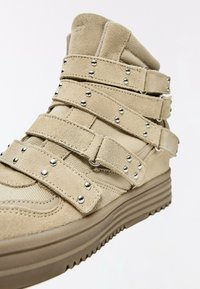 Beige high-top sneaker with suede and mesh materials, featuring multiple straps with metal studs and a thick ridged rubber sole.