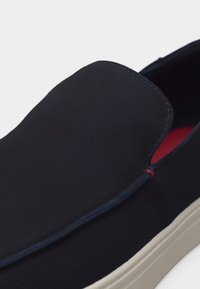 Μπλε ναυτικό casual παπούτσι τύπου slip-on με λευκή σόλα και κόκκινη εσωτερική επένδυση, παρουσιάζοντας κοντινή λήψη της μπροστινής επάνω και πλαϊνής ραφής.