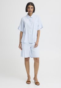 Femme aux cheveux courts et foncés portant une chemise à manches courtes bleu clair à boutons et un short assorti jusqu'aux genoux, talons ouverts marron, debout sur un fond blanc.