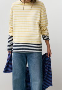 Personne portant des hauts à manches longues superposés à rayures jaunes et bleu marine, jean bleu, tenant un tissu rayé bleu et bordeaux.