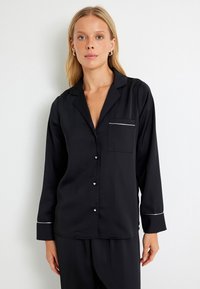 Zwarte satijnen pyjama-top met een reverskraag, een voorzak rand in het wit en knoopsluiting. Heeft lange mouwen en een gladde textuur.