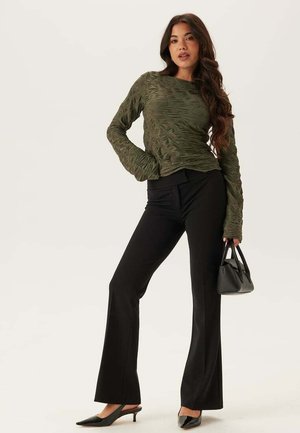 Kvinde iført struktureret olivengrøn langærmet top, sorte vide bukser, sorte slingback-hæle, holder en lille sort håndtaske, stående på hvid baggrund.